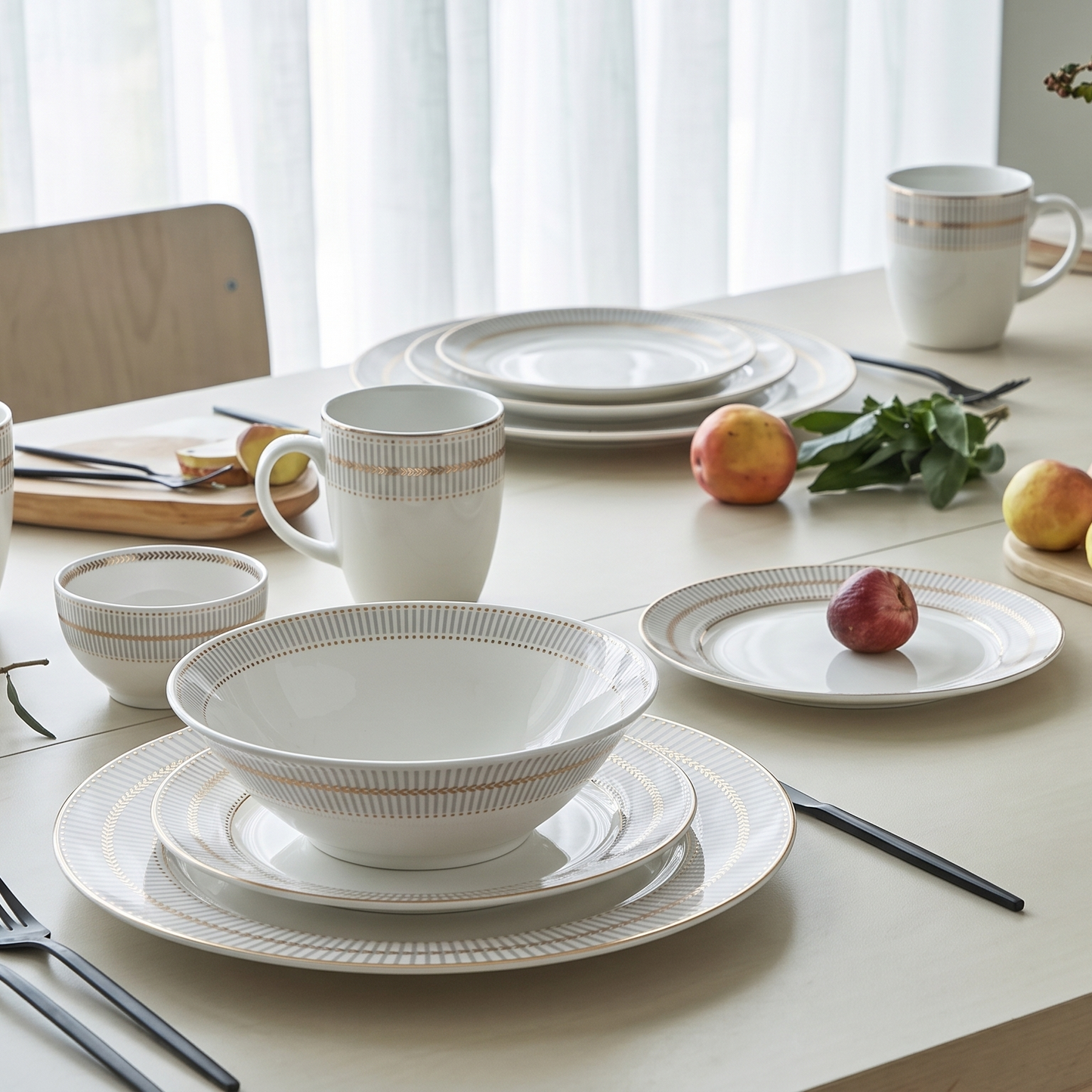 A styled dining table setting featuring a coordinated white ceramic tableware collection with subtle gold-beige striped detailing. The set includes plates, bowls, a mug, and forks, arranged on a light wood table with fruit, greenery, and chairs in the background.