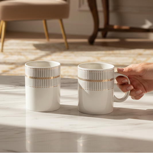 Two white ceramic mugs with gold and beige striped bands are shown on a marble surface in a sunlit room. One mug is standing alone, and a hand is holding the handle of the second mug.