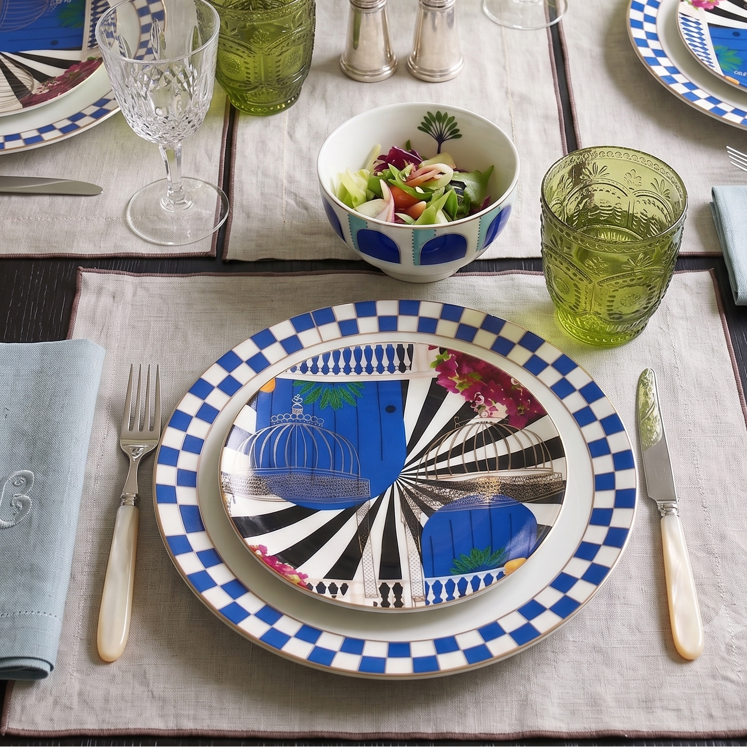 A styled table setting featuring matching blue and white patterned plates, green textured drinking glasses, light blue napkins, a floral centerpiece, and a bowl of salad on a beige placemat.