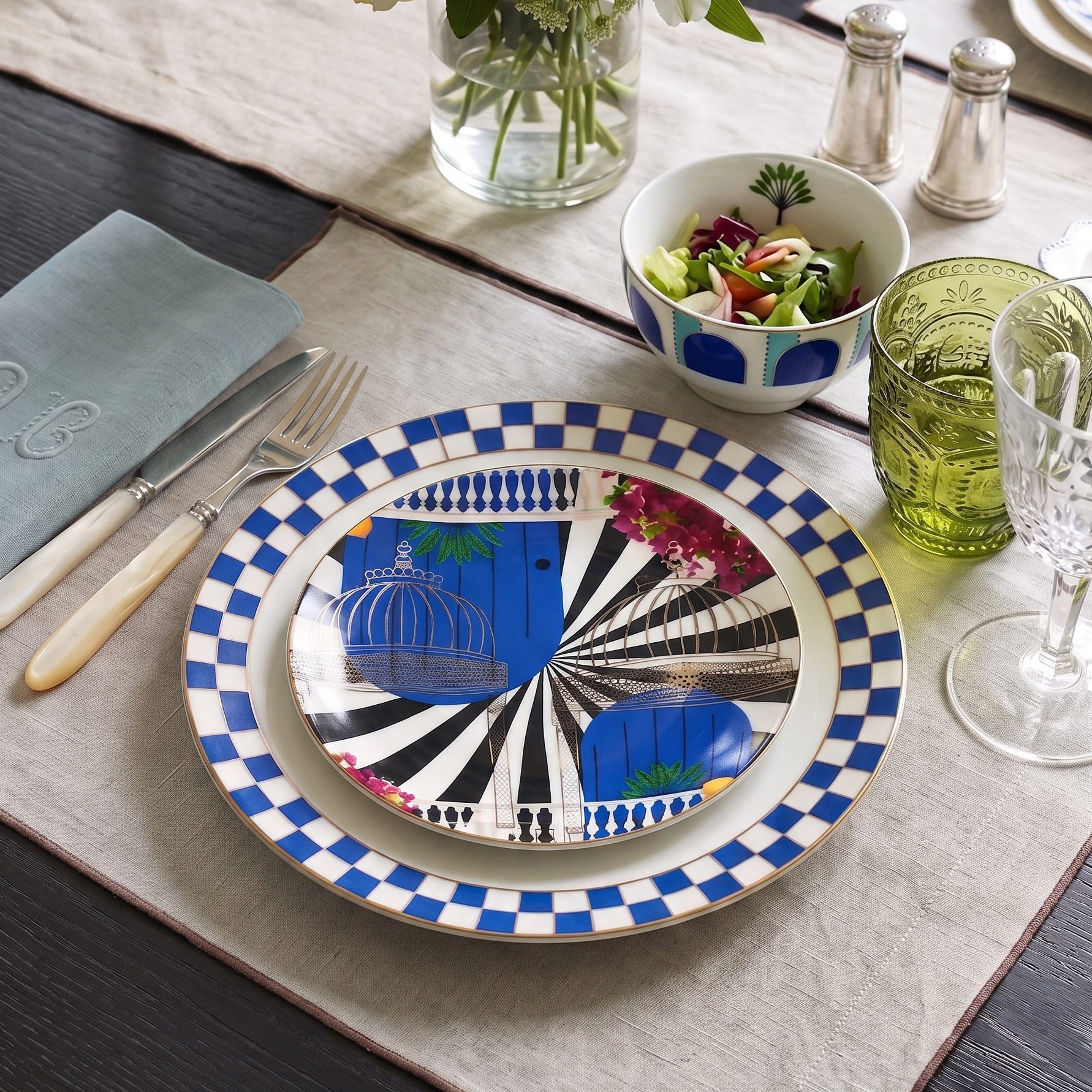A styled dining table setting featuring a round ceramic plate with a blue and white checkered rim and a central black-and-white striped design with a blue birdcage motif. The plate is set on a beige placemat with silverware, a glass vase of flowers, a bowl of salad, and green and clear drinking glasses.