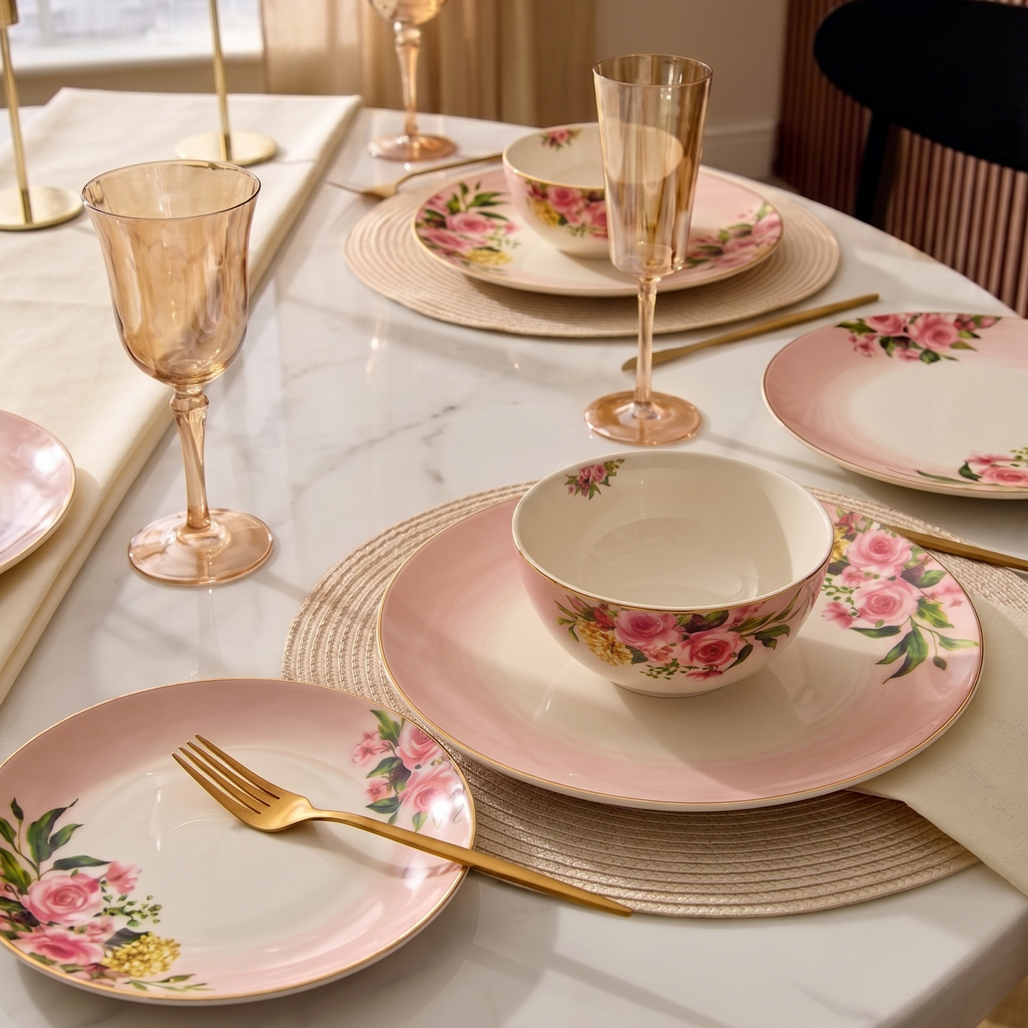 A styled dining table setting featuring a pink floral ceramic dinnerware set with matching bowls, plates, and gold-toned glassware on a white marble table runner.