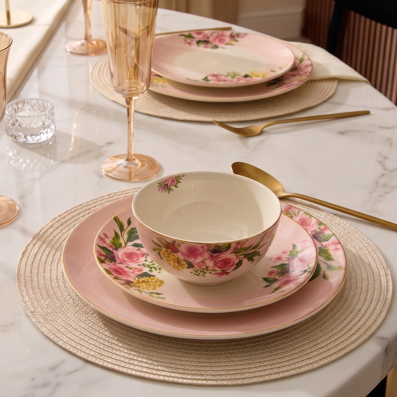 A styled dining table setting featuring a pink and white floral porcelain dinnerware set with matching pink plates, gold-toned cutlery, amber-tinted glass goblets, woven placemats, and a cream marble tabletop.