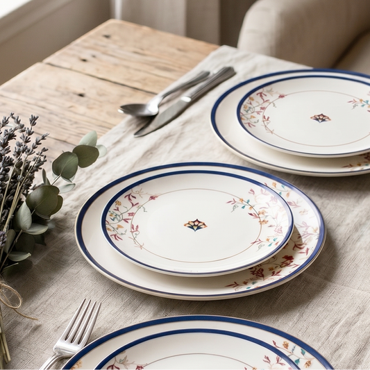 A set of white ceramic dinner plates with blue rims and delicate floral patterns, arranged on a rustic wooden table with silver cutlery, a linen tablecloth, and decorative greenery.